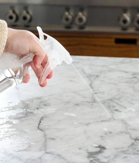 A hand spraying cleaner on a marble kitchen countertop, emphasizing regular maintenance for longevity.